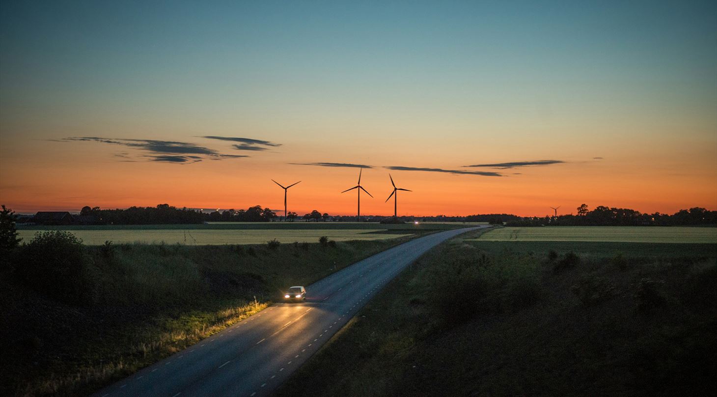 En bil krer ad en de landevej i skumringen mod en horisont, der er oplyst af en smuk solnedgang. Tre vindmller str silhouetteret mod den orange og bl himmel, omgivet af marker og trer.